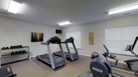 This fitness room offers a dedicated space for cardiovascular exercise, designed to meet everyday workout needs. The room features neutral gray walls and carpeted flooring, creating a clean and functional environment. Two treadmills are positioned as the focal point of the space, accompanied by an elliptical machine and a stability ball, providing a variety of cardio and balance training options. A wall-mounted flat-screen television is installed above eye level, allowing users to stay entertained during workouts. Natural light filters into the room through two windows fitted with white blinds, brightening the space alongside overhead fluorescent lighting. Decorative wall art adds a subtle visual touch, keeping the atmosphere from feeling purely utilitarian. The layout is straightforward and efficient, making good use of the available floor space without feeling cluttered. Overall, this fitness room provides a convenient and practical setting for residents or users looking to maintain an active lifestyle without leaving the property.