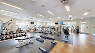 Located at 1200 Somerby Dr in Mobile, Alabama, this well-appointed fitness center spans a generous single-floor layout divided into two exercise rooms, offering a comprehensive environment for a wide range of workout routines. The facility is bright and open, with large windows lining the walls that flood the space with natural light and provide views of the surrounding greenery outdoors. Ceiling fans are positioned throughout to maintain comfortable airflow during workouts.

The main exercise room is expansively proportioned and features a robust selection of cardio equipment, including multiple treadmills, elliptical trainers, stair climbers, and stationary bikes. Flat-screen televisions are mounted at convenient viewing angles to keep users engaged during cardio sessions. A full-length wall mirror visually expands the space while supporting form checks during exercise. Rubber floor mats are laid across dedicated workout zones, complementing the warm terra-cotta tile flooring throughout.

The second exercise room provides a strength-training focus, housing a variety of resistance machines, a dumbbell rack with a graduated free-weight selection, weight benches, and a multi-use cable machine. Crown molding and recessed lighting add a refined architectural touch, giving the facility a polished, upscale ambiance. Together, both rooms provide a well-rounded, thoroughly equipped fitness experience.