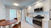 This well-appointed single-floor residence offers a thoughtfully designed layout that balances functionality and modern style. The kitchen is outfitted with crisp white cabinetry, white subway tile backsplash, light-toned countertops, and a full suite of stainless steel appliances including a gas range, over-the-range microwave, dishwasher, and full-size refrigerator. Light tile flooring ties the kitchen seamlessly into the adjacent dining area, which features a warm wood round table with hairpin legs and matching wood chairs, adding a mid-century modern touch to the space. A dark wood sliding entry door adds a striking contrast against the neutral walls. The bedroom is a standout feature, highlighted by expansive floor-to-ceiling windows that invite abundant natural light and offer open views of the surrounding urban streetscape. Light hardwood flooring, a platform bed with an integrated walnut headboard and built-in nightstands, and a concrete structural column lend the room both warmth and an industrial-modern character. The bathroom is finished with clean white rectangular wall tiles, a soaking tub with overhead shower, light stone-look flooring, and a mirrored medicine cabinet, creating a serene and polished retreat.