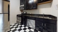 Located at 3408 Park Ave in Richmond, Virginia's 23221 zip code, this single-floor residence offers a well-organized layout with a cohesive blend of classic and functional design elements. Rich hardwood flooring flows throughout the main living and bedroom areas, lending warmth and character to each room. The living area welcomes you through an elegant arched entryway, complemented by neutral walls, crisp white trim, and a textured ceiling that adds subtle visual interest. The bedroom is generously sized, featuring multiple windows with adjustable blinds that invite ample natural light, a built-in shelving unit with sliding doors for storage, and direct access to an exterior door. The kitchen stands out with its striking black-and-white checkered tile floor, dark espresso cabinetry, granite countertops, and a full suite of stainless steel appliances including a gas range, built-in microwave, dishwasher, and sink. The bathroom includes a full soaking tub with tiled surround and is accessible via a classic paneled door. Throughout the space, consistent use of white interior doors with antique brass hardware ties the design together, creating a cohesive and inviting atmosphere suited for comfortable everyday living.