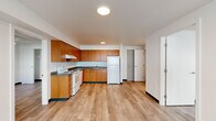 This 2-bedroom, 1-bathroom unit features a clean, modern design with light wood-style flooring extending throughout the main living areas and bedrooms. The open kitchen is equipped with warm-toned wood cabinetry, ample upper and lower storage, gray laminate countertops, a stainless steel sink, and white appliances including an electric range, dishwasher, and refrigerator — all laid out in an efficient L-shaped configuration that flows naturally into the living area.

The primary bedroom is a comfortable, well-proportioned room featuring a ceiling fan with integrated lighting, a window that brings in natural light, and a dedicated closet for storage. The second bedroom is similarly bright, with its own window overlooking the surrounding building exterior and light-toned flooring. A separate room features dual sliding-door closets with wire shelving, providing generous additional storage space.

The bathroom is clean and functional, finished with a wood-toned vanity, gray countertop, single-basin sink, a large mirror, and a full bathtub. Speckled tile flooring adds a practical touch to the space. Throughout the unit, dark baseboards contrast neatly with the white walls, contributing to a crisp, contemporary aesthetic. A connecting hallway ties all rooms together efficiently.