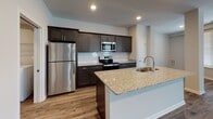This well-appointed single-floor residence offers a thoughtfully designed open-concept layout that seamlessly connects the main living and kitchen areas. The kitchen is a standout feature, showcasing rich dark cabinetry, granite countertops, a subway tile backsplash, and a full suite of stainless steel appliances including a refrigerator, range, and built-in microwave. A large center island with an undermount sink and additional seating provides both functionality and a natural gathering point.

The living area is generously proportioned, bathed in natural light from multiple windows fitted with blinds, and finished with durable hardwood-style flooring that extends throughout the main spaces. A dedicated hallway leads to the private areas of the residence, where bedrooms feature plush carpet flooring and bright windows. The primary bedroom connects to a private bathroom with a granite-topped dark vanity, tile flooring, and direct access to a spacious walk-in closet equipped with wire shelving. A second full bathroom mirrors the same design aesthetic, complete with a soaking tub and shower combination. A dedicated laundry room houses a full-size front-loading washer and dryer with overhead wire shelving, adding everyday convenience to this clean and modern home.