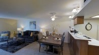This single-floor residence offers a thoughtfully designed open-concept layout that integrates the living, dining, and kitchen areas into one cohesive and functional space. The dining area serves as a central hub, accommodating a solid wood rectangular dining table with upholstered seating for up to six guests, illuminated by a multi-bulb ceiling light fixture that adds a warm, inviting glow. A decorative centerpiece with branching botanicals adds a natural, elegant touch to the table setting.

Adjacent to the dining space, the living room is furnished with a plush, dark charcoal sofa accented by mustard yellow throw pillows, paired with a soft area rug that defines the seating zone. A ceiling fan with a light fixture provides both airflow and ambient lighting overhead, while a tall floor lamp in the corner contributes to the warm, layered lighting throughout. Subtle wall art adds a splash of color and personality to the neutral-toned walls.

The kitchen transitions smoothly from the dining area, featuring dark cabinetry and a light countertop that offer a classic contrast. The overall ambiance of the space is warm and welcoming, with neutral tones, soft lighting, and well-chosen furnishings creating a comfortable and livable environment throughout.