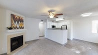 This single-floor residence offers a thoughtfully designed open-concept layout that connects the living area and kitchen in one cohesive space. The living room is highlighted by a classic fireplace with a tiled surround, adding warmth and character to the room. A ceiling fan with a light fixture is centrally positioned, promoting air circulation and providing ambient lighting. The carpeted flooring throughout the living area lends a comfortable, inviting feel. The kitchen is defined by a peninsula counter with a dark countertop that serves as both a workspace and a natural divider between the living and cooking areas. Crisp white cabinetry lines the kitchen walls, providing ample storage, while stainless steel appliances — including a refrigerator and microwave — add a modern touch. Under-cabinet lighting enhances the functionality of the workspace. A textured ceiling runs throughout the space, adding subtle visual depth. A hallway visible from the main area leads further into the residence, suggesting additional rooms beyond the open living zone. The overall aesthetic is clean, neutral, and move-in ready, with a bright and airy atmosphere created by the white walls and light-toned finishes.