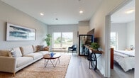 This thoughtfully designed single-floor residence offers a seamless blend of modern style and functional living. The open-concept layout connects the living room, dining area, and kitchen, creating an inviting and cohesive space throughout.

The living room is bright and welcoming, featuring large windows that frame expansive outdoor views of a green landscape. Light wood-style flooring, recessed lighting, and soft neutral wall tones lend a clean, contemporary feel to the space.

The kitchen is a standout feature, outfitted with sleek grey cabinetry extending to the ceiling, a full suite of stainless steel appliances including a refrigerator, range, and microwave, and a light stone countertop. A peninsula with seating for multiple bar stools serves as both a casual dining space and a natural gathering point, illuminated by elegant pendant lighting overhead.

The two bedrooms are comfortably proportioned, each with large windows offering views of the surrounding greenery. The rooms feature neutral carpet or hard flooring and ample wall space. The bathroom is clean and functional, with grey cabinetry, a vanity with under-sink storage, tile flooring, and a large mirror.

Throughout the residence, a cohesive palette of soft greys, warm wood tones, and natural textures creates a calm, modern ambiance.