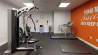Located at 735 Randolph St in Canton, Massachusetts, this single-room fitness center is designed to provide a functional and motivating workout environment. The space features a bold color scheme, with a vibrant orange accent wall serving as a focal point and displaying the motivational phrase "You Will Succeed, Just Keep Going!" — setting an energetic tone throughout.

The room is equipped with a variety of exercise machines, including a treadmill, an elliptical trainer, and a multi-station cable strength machine, offering options for both cardiovascular and resistance training. Durable rubber flooring covers the entire workout area, providing safety and cushioning, while exercise mats are available for floor-based workouts.

Overhead panel lighting ensures the space is well-lit, and a small window allows for natural light and outdoor views. A wall clock, a water dispenser, and a waste bin are thoughtfully included for convenience. Wall-mounted hooks offer storage for personal items. Safety features include a fire alarm system, an emergency exit door, and clearly posted signage.

The overall ambiance is clean, practical, and purpose-driven, making this fitness center a well-appointed resource for those seeking a straightforward and efficient place to exercise.