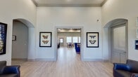 This single-story community clubhouse at Capella Apartment Homes is thoughtfully designed to serve as a central gathering space for residents. The entry foyer immediately impresses with its soaring ceilings and light-filled atmosphere, flanked by framed butterfly botanical prints that establish a cohesive decorative theme carried throughout the space. Light wood-look tile flooring flows seamlessly across all areas, contributing to a bright and airy aesthetic.

The welcoming lobby features a seating nook with a leather armchair and spindle bench, large windows admitting generous natural light, and a striking oversized butterfly artwork. A wide doorway connects this reception area to the main common spaces beyond.

The lounge area is anchored by a shiplap accent wall displaying a wall-mounted flat-screen television, floating wooden shelves with curated décor, and bold cobalt blue wingback chairs arranged around a colorful braided rug, creating a relaxed yet stylish gathering spot.

The fully equipped kitchen features dark espresso cabinetry with white countertops, a stainless steel side-by-side refrigerator, decorative columns, and dedicated coffee station. Adjacent dining areas offer multiple wooden tables with black chairs, abundant window light, and direct exterior access, making the space well-suited for community events and casual gatherings.
