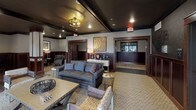 This thoughtfully designed single-floor communal space exudes a refined, upscale ambiance through its cohesive use of dark tones, rich wood accents, and carefully curated furnishings. The main lounge area serves as the centerpiece of the space, featuring plush charcoal leather sofas with nailhead trim, coordinating accent chairs, and a patterned area rug anchoring the seating arrangement. A textured dark ceiling with recessed lighting and an industrial-style flush-mount fixture adds depth and warmth overhead, while rich mahogany-toned wood trim frames doorways and wainscoting throughout. Distinctive wall art, including a striking wood-slice mosaic piece, contributes to the space's eclectic yet polished character. An additional lounge nook with booth-style seating and a framed artwork provides an intimate retreat within the larger room. The wet bar area is a standout feature, finished with dark cabinetry, dramatic granite countertops with bold veining, a stone tile backsplash, and a warm butcher block bar top. Pendant lighting, a wall-mounted television, and a mirrored accent panel complete the bar's functional and stylish design. Hardwood flooring transitions seamlessly throughout, tying the space together with a sense of continuity and sophistication.