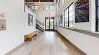 Located at 201 N Third St in Allentown, Pennsylvania, this two-floor lobby makes a striking first impression through its thoughtful blend of industrial and contemporary design. The space spans two levels, with the main floor commanding the majority of the footprint and offering an expansive, open feel enhanced by dramatically soaring ceilings.

Large, black-framed industrial windows line the walls, flooding the interiors with generous natural light and offering views to the surrounding streetscape. Light wood-look flooring runs throughout, providing a warm contrast to the crisp white walls and exposed ductwork above. A cascading arrangement of Edison bulb pendant lights descends from the ceiling, serving as a distinctive design focal point that bridges raw industrial character with modern elegance.

The main seating area is thoughtfully appointed with plush velvet armchairs, a contemporary sofa, and a round glass-top coffee table set atop a woven area rug — creating an inviting lounge atmosphere. Curated wall art, including a large colorful canvas and a sculptural textile piece, add artistic character throughout. A rustic reclaimed wood bench anchors the entry corridor near the glass double doors, complementing the space's mix of textures. An elevator and staircase with metal railings provide access between floors, completing this well-designed, welcoming lobby.