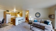 This single-floor residence showcases an open-concept design that seamlessly integrates the kitchen and living area into one cohesive space. The living room is carpeted and comfortably sized, centered around a neutral-toned sofa accented with a teal throw pillow, a dark-wood coffee table, and a side table with a classic lamp. A decorative round mirror with a textured metallic frame serves as a focal point on the main wall, adding visual interest to the otherwise clean, light-colored walls.

The kitchen is positioned adjacent to the living area and is separated by a raised breakfast bar with two bar stools, providing both a casual dining option and a natural dividing element between spaces. The kitchen features warm wood-toned cabinetry, a countertop workspace, and under-cabinet lighting that adds warmth to the cooking area. A trailing plant atop the upper cabinets introduces a touch of greenery.

A hallway visible beyond the kitchen leads to additional rooms, suggesting a thoughtful and functional floor plan. Overall, the space maintains a warm, neutral palette with practical furnishings and a welcoming ambiance suited for everyday living.