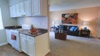 This single-floor residence offers a practical and welcoming layout designed to maximize comfort and functionality. The kitchen is outfitted with white cabinetry, dark countertops, and a gas range, providing ample storage and workspace for meal preparation. A ventilation hood overhead and clean, neutral wall tones give the kitchen a crisp, organized feel. The kitchen flows seamlessly into the living area through an open pass-through design, creating a connected and sociable atmosphere. The living room features plush carpeting underfoot, offering a warm contrast to the hard flooring of the kitchen. The space is generously proportioned, easily accommodating a full sofa and additional seating, along with accent furniture such as a side table and ottoman. Soft, neutral wall tones throughout the unit create a calm, versatile backdrop that complements a variety of décor styles. Ambient lighting contributes to the cozy ambiance of the living area. Overall, the layout reflects a thoughtful single-floor design that balances open connectivity between spaces with a sense of defined, purposeful living areas — ideal for those seeking a comfortable and easy-to-navigate home.