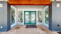 The Paxton leasing office is a thoughtfully designed single-story space that makes an immediate impression with its striking exterior entrance — bold teal French doors framed by large sidelight windows set beneath warm exposed wood beam ceilings, creating a welcoming first arrival experience. Inside, the open-plan interior is finished with large-format beige tile flooring that runs seamlessly throughout, lending a clean, cohesive aesthetic to the entire space. A standout feature is the central kitchen and bar area, highlighted by a substantial island clad in stacked natural stone with a light granite countertop, dark cabinetry, a stone tile backsplash, and stainless steel appliances — offering a functional yet visually rich focal point. Adjacent to the kitchen, a high-top dining area provides an inviting spot for casual seating, bathed in natural light from oversized windows with views to the surrounding community. The lounge area features a contemporary sofa, colorful artwork, and generous window coverage that keeps the space bright and airy. A rustic sliding barn door adds a charming contrast against the otherwise modern finishes, leading to a secondary utility area. Throughout the office, branded Paxton rugs and signage reinforce the community identity, while recessed lighting and ample windows ensure the space feels open and well-lit at all times.