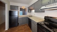 This single-floor space is designed with functionality in mind, centered around a well-equipped kitchen that serves as the heart of the layout. The kitchen features gray cabinetry spanning both upper and lower sections, providing ample storage throughout. Light-toned laminate countertops offer generous workspace, complemented by a stainless steel sink, a black electric range with a ventilation hood above, a black dishwasher, and a full-size black refrigerator-freezer. An under-cabinet fluorescent light fixture adds task lighting to the prep area. The kitchen opens through a pass-through window that connects to an adjoining area, enhancing the sense of openness. Wood-look flooring runs through the kitchen and into the connecting hallway, adding warmth to the space. The walls throughout feature a textured finish in neutral tones. A secondary room offers a more intimate setting, with wood-look flooring, a window fitted with horizontal blinds that allows natural light to filter in, a floor vent for climate control, and a textured wall finish consistent with the rest of the space. The overall layout is compact and practical, with clear sightlines between areas.