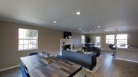 Located in Denton, this single-story space offers a thoughtfully designed open-concept layout that seamlessly connects the living and dining areas. The main living space is anchored by a classic white-surround fireplace, adding a focal point of warmth and elegance to the room. Recessed lighting is installed throughout the ceiling, complementing the natural light that pours in through multiple windows along the walls. Rich hardwood flooring extends across the space, providing a warm and cohesive aesthetic underfoot. The dining area features a sturdy wooden table that pairs well with the overall neutral color palette of warm taupe walls and white crown molding trim. The living area is furnished with contemporary dark upholstered seating arranged around a patterned area rug, creating an inviting and functional gathering space. A wall-mounted flat-screen television is positioned above the fireplace for comfortable viewing. A glass-paneled door at the rear of the space allows additional natural light to filter in while providing access to the exterior. The combination of classic architectural details, modern finishes, and an open floor plan makes this space both practical and visually appealing.