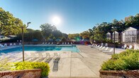 Located at 1200 Trillium Cir in Raleigh, NC, this expansive outdoor recreational facility offers a resort-style pool experience set within a beautifully maintained, tree-lined environment. The centerpiece of the space is a large, rectangular swimming pool with crystal-clear turquoise water, accessible via multiple entry points including handrail-equipped steps and a dedicated shallow end marked at 3 feet. The pool is surrounded by a generous concrete sun deck that accommodates an extensive array of lounge chairs and Adirondack-style seating, providing ample space for sunbathing and relaxation. Vibrant blue patio umbrellas are positioned throughout the deck, offering shaded retreats on sunny days. A support building with modern white vertical board-and-batten siding and a sloped roof is situated adjacent to the pool area, accessible via a broad brick-lined staircase flanked by lush landscaping. Brick perimeter walls define the boundary of the facility, adding a classic architectural character to the space. Tall, mature trees encircle the entire area, creating a natural, private backdrop and providing a sense of seclusion. The overall ambiance is bright, open, and welcoming, ideal for community recreation and leisure in the heart of Raleigh.