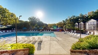Located at 1200 Trillium Cir in Raleigh, NC, this expansive outdoor recreational facility offers a resort-style pool experience set within a beautifully maintained, tree-lined environment. The centerpiece of the space is a large, rectangular swimming pool with crystal-clear turquoise water, accessible via multiple entry points including handrail-equipped steps and a dedicated shallow end marked at 3 feet. The pool is surrounded by a generous concrete sun deck that accommodates an extensive array of lounge chairs and Adirondack-style seating, providing ample space for sunbathing and relaxation. Vibrant blue patio umbrellas are positioned throughout the deck, offering shaded retreats on sunny days. A support building with modern white vertical board-and-batten siding and a sloped roof is situated adjacent to the pool area, accessible via a broad brick-lined staircase flanked by lush landscaping. Brick perimeter walls define the boundary of the facility, adding a classic architectural character to the space. Tall, mature trees encircle the entire area, creating a natural, private backdrop and providing a sense of seclusion. The overall ambiance is bright, open, and welcoming, ideal for community recreation and leisure in the heart of Raleigh.