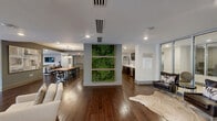 Located at 6205 Carnegie Blvd in Charlotte, NC, this generously sized single-floor amenity space is thoughtfully designed to accommodate both social gatherings and everyday use. Rich hardwood flooring flows seamlessly throughout, anchoring the modern yet welcoming aesthetic defined by neutral tones, recessed lighting, and tray ceilings.

The fully equipped kitchen is a standout feature, offering a galley-style layout with dark cabinetry, crisp white quartz countertops, stainless steel appliances including a professional-grade gas cooktop, a large range hood, and a full-size refrigerator. A bold geometric-patterned tile backsplash adds a stylish focal point to the space.

Two distinct dining areas serve different purposes: one features a live-edge wood communal table beneath a statement chandelier, ideal for larger gatherings, while the other offers a more intimate setting with a gray-toned rectangular table, elegant white chairs, and a decorative pendant light overhead.

The living area provides a comfortable lounge environment with multiple seating arrangements, built-in media consoles, wall-mounted televisions, and large windows that invite natural light and outdoor views. A glazed double-door entry connects the space to an adjacent outdoor courtyard area, enhancing the open, airy atmosphere throughout.