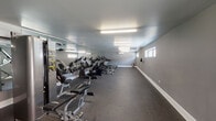 This fitness center offers a comprehensive and thoughtfully designed workout environment spread across a single floor. The space features a neutral grey color palette throughout, with white baseboards and a smooth ceiling outfitted with bright fluorescent lighting that ensures excellent visibility across the entire floor.

The main workout area is generously sized and lined with full-wall mirrors along one side, creating an open and expansive feel while allowing users to monitor their form during exercise. The durable rubber flooring provides both safety and comfort underfoot. A row of cardio equipment — including elliptical trainers, stationary bikes, and treadmills — runs the length of the mirrored wall, offering a well-stocked selection for aerobic training.

Toward one end of the room, a multi-station strength machine and an adjustable weight bench provide options for resistance and strength training. A wall-mounted split A/C unit ensures climate control throughout the space. Natural light filters in through several horizontally positioned windows positioned high along the exterior walls, complementing the artificial lighting.

Additional amenities include a wall-mounted water fountain, a waste bin, and clearly marked exits, contributing to the space's practical and user-friendly design. The overall ambiance is clean, functional, and well-maintained — making it a capable facility for a full range of fitness activities.
