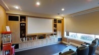 This single-floor media room is thoughtfully designed to deliver a premium home theater experience. The focal point of the space is a large projection screen seamlessly integrated into an expansive wall of custom built-in cabinetry. The cabinetry features a combination of open shelving — generously stocked with books and media collections — alongside closed cabinet storage below, providing ample organization while maintaining a clean, structured aesthetic.

The warm, neutral tones of the cabinetry complement the soft yellow walls and smooth white ceiling, creating an inviting and cozy atmosphere. Recessed lighting is evenly distributed across the ceiling, offering controlled, adjustable illumination ideal for both viewing and casual use. Dark hardwood flooring anchors the room with a rich contrast against the lighter furnishings.

A window fitted with honeycomb roller blinds along the side wall allows for natural light when desired while also enabling full light control for optimal screen visibility during viewing sessions. The presence of comfortable recliner seating enhances the cinematic feel of the room. Overall, this space strikes a well-balanced blend of functional entertainment design and warm residential comfort, making it equally suited for movie nights, gaming, or quiet reading.