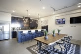 This single-floor space makes a strong design statement through a carefully curated blend of bold color, modern fixtures, and thoughtful functionality. The kitchen is a standout centerpiece, showcasing floor-to-ceiling navy blue cabinetry with gold hardware, crisp white quartz countertops, and a mosaic tile backsplash that adds texture and visual depth. A large island with bar seating runs along one side, topped with a sculptural multi-bulb chandelier that serves as an artistic focal point. Premium stainless steel appliances, including a professional-grade range with a wall-mounted hood, a built-in microwave, and a panel-ready refrigerator, round out the culinary suite. Light tile flooring runs throughout, keeping the space feeling open and cohesive. The dining area is equally distinctive, anchored by a live-edge wood table paired with sleek black wishbone chairs atop a bold black-and-white geometric tile inlay. A full-width glass panel garage door opens the dining space to the outdoors, flooding the room with natural light and creating a seamless indoor-outdoor connection. A wall-mounted television and recessed lighting add practical comfort, while the overall design delivers a contemporary, sophisticated ambiance throughout.