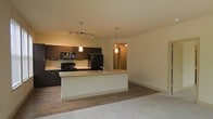 This single-floor space showcases a thoughtfully designed open-concept layout that seamlessly connects the kitchen and living area. The kitchen is appointed with rich, dark cabinetry that provides a striking contrast against the light-toned countertops and walls. A central peninsula with an integrated sink serves as both a functional workspace and a natural divider between the kitchen and living area, offering additional seating and entertaining space. Black appliances, including a range, microwave, and refrigerator, lend a sleek, cohesive look to the kitchen. Overhead, a multi-head track lighting fixture illuminates the cooking area, while two elegant pendant lights hang above the peninsula, adding warmth and ambiance. The flooring transitions from rich hardwood-style planks in the kitchen and dining zone to plush carpet in the living area, defining each space while maintaining an open feel. Neutral, warm-toned walls throughout contribute to a welcoming atmosphere. A hallway visible in the background suggests additional rooms beyond, further enhancing the functional layout of this well-appointed space.