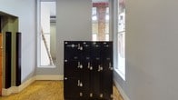 This single-floor Laundry Locker facility is a purpose-built, self-service laundry solution designed for convenience and accessibility. The space is entered through a distinctive purple door, opening into a clean, functional area with light gray walls and composite wood flooring throughout. The room is well-lit by large windows that flood the interior with natural light, framing views of exposed brick elements that hint at the building's historic character.

At the heart of the facility stands a bank of sleek, black Laundry Locker units. These secure, individually keyed lockers are clearly labeled and organized for straightforward use, accommodating drop-off and dry-cleaning services. The dark finish of the lockers provides a strong visual contrast against the neutral walls and bright surroundings.

A prominent feature of the space is the industrial orange freight elevator, which serves as the primary means of access and is designated for operation during set hours. Protective black bumper guards flank the elevator entrance, emphasizing its utilitarian role. An emergency lighting fixture is also present, ensuring safety within the space.

Overall, the facility maintains a practical, no-frills ambiance while offering a well-organized and clearly functional service environment tailored to efficient laundry management.