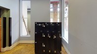 This single-floor Laundry Locker facility is a purpose-built, self-service laundry solution designed for convenience and accessibility. The space is entered through a distinctive purple door, opening into a clean, functional area with light gray walls and composite wood flooring throughout. The room is well-lit by large windows that flood the interior with natural light, framing views of exposed brick elements that hint at the building's historic character.

At the heart of the facility stands a bank of sleek, black Laundry Locker units. These secure, individually keyed lockers are clearly labeled and organized for straightforward use, accommodating drop-off and dry-cleaning services. The dark finish of the lockers provides a strong visual contrast against the neutral walls and bright surroundings.

A prominent feature of the space is the industrial orange freight elevator, which serves as the primary means of access and is designated for operation during set hours. Protective black bumper guards flank the elevator entrance, emphasizing its utilitarian role. An emergency lighting fixture is also present, ensuring safety within the space.

Overall, the facility maintains a practical, no-frills ambiance while offering a well-organized and clearly functional service environment tailored to efficient laundry management.