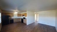 This single-story space showcases an open-concept layout that seamlessly connects the kitchen and living area, creating a cohesive and functional environment. The kitchen features light oak cabinetry with dark granite-style countertops, providing ample storage and workspace. Black appliances, including a range and dishwasher, are integrated into the design, complemented by an under-cabinet range hood. A ceiling fan with a light fixture is centrally positioned, ensuring comfortable airflow throughout the shared space. The continuous dark wood-style flooring flows throughout, adding warmth and visual consistency to the interior. Neutral white walls keep the atmosphere bright and versatile, offering a clean canvas for various décor styles. A half-wall partition subtly defines the kitchen from the open living space without disrupting the airy feel of the layout. A doorway visible from the main area leads to additional rooms beyond, suggesting further living quarters. The overall design is practical and straightforward, with a focus on easy maintenance and an adaptable floor plan suited to a variety of lifestyles.