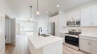 This well-designed single-story home offers three bedrooms and two bathrooms across a thoughtfully laid-out floor plan. The open-concept kitchen and living area serves as the heart of the home, anchored by a large central island with a white quartz countertop, an undermount sink with a matte black faucet, and stylish pendant lighting overhead. The kitchen features a full suite of stainless steel appliances, including a refrigerator, range, and microwave, complemented by white shaker-style upper cabinets, grey lower cabinetry, and generous counter space. A walk-in pantry provides additional storage. The living room is bright and airy, with light wood-look flooring, a ceiling fan, and large windows that invite natural light. Sliding glass doors open to a private balcony with scenic views of surrounding trees. The bedrooms are carpeted for comfort, offering a cozy retreat. The primary bathroom includes a walk-in shower with a glass enclosure, a white vanity with quartz countertop, and large-format tile flooring. The secondary bathroom mirrors this clean aesthetic with a white vanity, tile flooring, and modern fixtures. Throughout, the home maintains a crisp, neutral palette that creates a bright and welcoming ambiance.