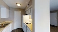 This single-story space showcases a functional kitchen area designed for efficient use of space. The kitchen is equipped with a white electric coil-top range and oven, a full-size refrigerator, and overhead cabinetry that provides practical storage. Light-toned countertops complement the white appliances, while an under-cabinet light adds focused illumination to the cooking area. A ceiling-mounted flush light fixture provides general lighting throughout the space. The walls are finished in a warm taupe tone, paired with a textured ceiling that adds subtle depth to the interior. Dark hardwood-style flooring runs through the kitchen and into the adjacent hallway, creating a cohesive visual flow. A ceiling vent ensures proper climate control throughout the unit. A white interior door visible at the end of the hallway leads to an additional area of the space. The overall design is straightforward and practical, with a neutral color palette that offers a clean, understated aesthetic suitable for everyday living.