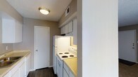 This single-story space showcases a functional kitchen area designed for efficient use of space. The kitchen is equipped with a white electric coil-top range and oven, a full-size refrigerator, and overhead cabinetry that provides practical storage. Light-toned countertops complement the white appliances, while an under-cabinet light adds focused illumination to the cooking area. A ceiling-mounted flush light fixture provides general lighting throughout the space. The walls are finished in a warm taupe tone, paired with a textured ceiling that adds subtle depth to the interior. Dark hardwood-style flooring runs through the kitchen and into the adjacent hallway, creating a cohesive visual flow. A ceiling vent ensures proper climate control throughout the unit. A white interior door visible at the end of the hallway leads to an additional area of the space. The overall design is straightforward and practical, with a neutral color palette that offers a clean, understated aesthetic suitable for everyday living.