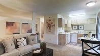 This single-floor unit showcases an open-concept design that seamlessly connects the living area and kitchen, creating a spacious and functional flow. The living area is carpeted and features a textured ceiling, neutral walls adorned with decorative artwork, and enough room to accommodate a comfortable seating arrangement centered around a coffee table. Natural and overhead lighting contribute to the warm, inviting ambiance of the space.

The kitchen is positioned adjacent to the living area and is outfitted with wood-toned cabinetry providing ample storage, white appliances including a range and dishwasher, and a clean countertop workspace. Upper and lower cabinets are thoughtfully arranged to maximize utility. A decorative floral arrangement displayed on the half-wall partition adds a welcoming touch between the two spaces.

A hallway visible from the main living area leads to a bathroom, suggesting additional private spaces beyond the open layout. The consistent use of neutral tones — beige, white, and warm wood accents — throughout the unit creates a cohesive and understated aesthetic, making the space feel bright, clean, and move-in ready.