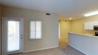 Located at 1390 Santa Alicia in Chula Vista, this single-story unit offers a straightforward, functional layout designed for comfortable everyday living. The living area features wall-to-wall carpeting in a neutral tone, contributing to a warm and understated aesthetic throughout the space. Warm beige walls provide a clean, versatile backdrop that complements a variety of furnishing styles.

A window fitted with white horizontal blinds allows natural light to filter into the living area, brightening the space while offering privacy control. Overhead, a centrally placed HVAC vent indicates a climate control system integrated into the ceiling, ensuring year-round comfort. Recessed lighting in the adjoining area provides additional illumination, enhancing the sense of depth and continuity between rooms.

Toward the rear of the living space, a partial wall and raised countertop define the transition into the kitchen area, creating a semi-open layout that maintains separation while preserving an open feel. White baseboards frame the perimeter of the rooms, adding a crisp, finished detail to the overall design. The unit presents a clean, move-in ready condition well-suited for a range of residents.
