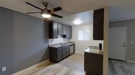 This single-floor space offers a clean, modern aesthetic with thoughtful design details throughout. The kitchen is a standout feature, showcasing dark espresso-toned cabinetry that extends to the ceiling for ample storage, complemented by granite-style countertops and a stainless steel dishwasher. A double-basin sink sits beneath a window fitted with vertical blinds, allowing natural light to filter into the space. A peninsula counter provides additional prep space and a casual dining option. The light wood-style flooring runs seamlessly throughout, creating a warm and cohesive look that contrasts beautifully with the dark cabinetry. An accent wall painted in a cool gray tone adds depth and a contemporary touch to the open layout. The textured ceiling features a ceiling fan with an integrated light fixture, ensuring comfortable airflow and functional lighting. Flush-mount ceiling lights provide additional illumination. The overall ambiance is clean, functional, and modern, making excellent use of the available space while maintaining a stylish and inviting atmosphere suitable for everyday living.