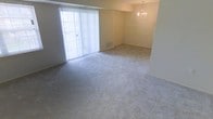 This single-floor space features an open layout with wall-to-wall carpeting throughout, creating a cohesive and comfortable feel across the living areas. A large sliding glass door fitted with vertical blinds anchors one wall, allowing generous natural light to fill the space while providing access to the outdoors. The neutral, beige-toned walls and light carpet complement each other, contributing to a bright and airy atmosphere. A stepped ceiling detail separates two connected living zones, with a decorative chandelier suspended in the adjoining area, adding a touch of elegance to the otherwise understated interior. A floor-level vent is visible along the baseboard, indicating a built-in climate control system. The open arrangement between the two areas offers flexible use of the space, suitable for both lounging and dining configurations. Overall, the space presents a clean, move-in-ready environment with classic, neutral finishes.