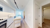 This single-floor unit showcases a clean, contemporary design throughout. The open-plan living and kitchen area is anchored by light hardwood flooring that extends seamlessly through the space, complemented by crisp white walls that enhance the sense of brightness. Track lighting runs along the kitchen corridor ceiling, providing functional illumination across the main living zone.

The kitchen is outfitted with modern stainless steel appliances, including a Blomberg refrigerator, built-in oven, microwave, and an induction cooktop. White flat-front cabinetry, white quartz countertops, and a textured tile backsplash complete the polished aesthetic. A freestanding white utility island adds versatile prep and storage space.

The living area benefits from large windows that frame urban views of the surrounding cityscape, allowing natural light to fill the room. Additional rooms offer neutral tones and light hardwood floors, including a smaller secondary space suitable for various uses.

The bathroom features a long vanity with warm wood cabinetry, dual sinks set in white countertops, a large mirror with sconce lighting, and tile flooring. A smart thermostat and electrical panel access are also present, reflecting thoughtful, modern infrastructure throughout the unit.
