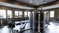 This fitness room offers a focused and well-appointed space for exercise, designed with both functionality and style in mind. The room is finished with deep, dark-toned walls that lend a sleek, modern aesthetic, complemented by dark hardwood-style flooring that ties the space together cohesively. Recessed lighting is strategically placed throughout the ceiling to ensure even illumination across the workout area, while a ceiling fan provides additional air circulation during intense sessions.

A row of treadmills lines one wall, each equipped with individual display consoles, positioned in front of a series of windows that allow natural light to filter into the space, creating an open and energizing environment. A large mirror on one wall enhances the sense of depth and allows users to monitor their form during workouts.

A multi-functional cable and weight machine occupies a prominent position in the room, providing options for a variety of strength training exercises. The overall layout is thoughtfully organized to maximize usable floor space while keeping equipment accessible and easy to navigate.