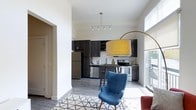 This single-floor space showcases a thoughtfully designed open-concept layout that seamlessly connects the living and kitchen areas. The soaring ceilings create a sense of spaciousness and airiness throughout, while large windows along the exterior wall flood the interior with abundant natural light.

The kitchen is outfitted with dark espresso-toned cabinetry that extends to the upper reaches of the tall walls, providing generous storage. Stainless steel appliances, including a refrigerator, range, and dishwasher, complement the contemporary aesthetic. A white subway tile backsplash adds a clean contrast to the dark cabinetry, and a small breakfast bar with seating offers a casual dining option.

The living area flows naturally from the kitchen, unified by light wood-toned flooring throughout. Crisp white walls serve as a neutral backdrop, allowing the architectural details and natural light to take center stage. A wide archway connects to an adjacent hallway, adding a subtle architectural element to the overall design. The open layout and generous ceiling height combine to give the space a welcoming, modern ambiance.