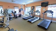 This single-floor fitness center offers a practical and comfortable environment for residents or members to maintain an active lifestyle. The room is finished with warm peach-toned walls that create an inviting atmosphere, complemented by carpeted flooring that adds a degree of comfort underfoot.

The space is outfitted with a selection of cardio equipment, including multiple treadmills positioned to make efficient use of the room's layout, as well as upright and recumbent stationary bikes for low-impact workout options. A wall-mounted flat-screen television is positioned for easy viewing during workouts, enhancing the overall exercise experience.

Natural light filters into the room through several windows dressed with decorative valances, providing a bright and pleasant setting. A glass-paneled door at the rear of the space allows additional light to enter and offers a connection to the outdoors. A water cooler is conveniently positioned near the entrance, ensuring hydration is easily accessible. Overhead fluorescent lighting supplements the natural light, keeping the space well-lit at all hours. A wall clock allows users to manage their workout time effectively. The overall layout is clean and organized, making this fitness center a functional and welcoming facility.
