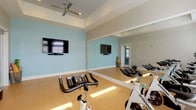 This single-floor spin studio is thoughtfully designed to support an engaging indoor cycling experience. The room is finished with smooth light wood flooring that reflects natural light streaming in through a row of large windows fitted with white horizontal blinds, offering views of outdoor greenery. The elevated tray ceiling, accented by a ceiling fan with integrated lighting, adds a sense of height and airiness to the space.

A full wall of floor-to-ceiling mirrors lines one side of the room, visually expanding the space and allowing cyclists to monitor their form during workouts. A wall-mounted flat-screen television paired with a sound bar is positioned on a soft aqua-blue accent wall, providing entertainment or instructor-led content during sessions. The remaining walls are painted in a light gray tone, contributing to the studio's clean, calm aesthetic.

The studio is equipped with a row of Spinning-brand stationary bikes arranged to face both the mirror wall and the windows, creating an energizing and functional layout. Yoga mats and accessories stored in a floor basket near the TV wall suggest the space also accommodates stretching or supplementary fitness activities. Overall, the studio combines practical fitness amenities with a bright, welcoming atmosphere.