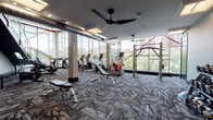 Located at 1350 Speer Boulevard in Denver, Colorado, this two-floor fitness center is a well-appointed facility designed to support a wide range of workout routines. The space is defined by its dramatic floor-to-ceiling glass walls that flood the interior with natural light and offer sweeping views of the Denver skyline and surrounding tree-lined streets.

The upper level serves as the primary cardio and strength training floor, featuring patterned carpet tiles and an array of machines including treadmills, ellipticals, stationary bikes, and cable machines. Exposed ductwork and ceiling fans add an industrial character to the space, while the expansive windows create an energizing, open atmosphere.

The lower level continues the fitness offering with a dedicated free weights and functional training area, complete with medicine ball racks, benches, and multi-use cable systems. An adjacent spinning studio features dark hardwood floors, wall-mounted televisions, and natural light from large windows. A separate studio space with mirrored walls and hardwood flooring provides ample room for group fitness classes or personal training sessions.

Throughout both floors, the design balances industrial aesthetics — exposed ceilings, bold linear lighting, and dark trim — with modern, functional fitness equipment, creating a dynamic and motivating environment for users.