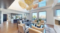 Located at 915 Thayer Ave in the heart of Silver Spring, MD, this elegantly designed amenity space spans a generous single floor and offers a seamless blend of luxury, function, and modern aesthetic. The centerpiece is a dramatic open-plan living area with soaring double-height ceilings, floor-to-ceiling windows that flood the space with natural light, and a stunning sculptural chandelier overhead. A stone-clad fireplace with a mounted TV anchors one wall, while rich hardwood flooring flows throughout. The gourmet kitchen features sleek, dark wood cabinetry, stainless steel appliances, and a large island, all framed by expansive windows overlooking the cityscape. A dedicated private dining room impresses with a large dark wood conference-style table, statement lighting, bold marble-patterned drapery, and city views beyond floor-to-ceiling glass doors that open to a terrace. The game room offers a full-size billiards table, patterned carpet, and direct outdoor access through sheer-curtained glass walls. The sprawling rooftop terrace is a standout feature, furnished with multiple seating arrangements and dining sets, surrounded by lush planters and railing views of the Silver Spring skyline.