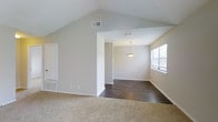 This single-floor residence offers a thoughtfully designed open-concept layout that seamlessly connects the living and dining areas. The living space is anchored by striking vaulted ceilings that soar overhead, creating a sense of volume and airiness throughout the room. Neutral, light-toned walls complement the space and contribute to its bright, welcoming ambiance.

The flooring transitions from plush, beige carpeting in the living area to rich, dark hardwood in the dining space, providing a clear yet fluid delineation between the two zones. A single pendant light fixture hangs above the dining area, adding a subtle decorative focal point. Natural light filters in through a window dressed with horizontal blinds, further enhancing the open and airy feel of the space.

Ceiling vents are strategically placed throughout, ensuring efficient climate control across the home. The clean, minimalist aesthetic is reinforced by crisp white trim and baseboards that frame each room. The overall design is practical and versatile, offering a neutral palette that serves as an ideal canvas for a variety of interior styles. This well-maintained, move-in ready home combines functional design with comfortable living in a single, cohesive floor plan.