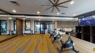 Located in Smyrna, TN, this well-appointed fitness center offers a comprehensive, multi-zone layout designed to support a wide range of workout routines. The facility is organized across distinct areas, beginning with a bright cycling studio equipped with multiple stationary bikes, a wall-mounted flat-screen TV, and full-length mirrors that amplify the sense of space. Striking striped carpet in bold yellow and grey tones runs throughout this zone, complemented by a large overhead ceiling fan that promotes air circulation during intense sessions.

Glass-paned double doors with black framing connect the cycling area to a dedicated strength and cardio room. This space features bold terracotta-orange accent walls, exposed ductwork lending an industrial aesthetic, and large windows that allow natural light to pour in while offering outdoor views. The room is outfitted with treadmills, ellipticals, rowing machines, weight benches, free weights, medicine balls, and a punching bag station, providing a thorough range of training options.

An adjacent private lounge or media room with a wall-mounted TV offers a space to cool down or stretch. The entrance area includes a body weight scale and informational signage. Throughout, the design balances modern aesthetics with functional fitness infrastructure, creating an energetic and well-equipped environment.