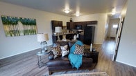 This two-floor residential space offers a thoughtfully designed open-concept layout that seamlessly connects the living and kitchen areas on the main level. The living area features warm wood-style laminate flooring that extends throughout, creating a cohesive and inviting atmosphere. The space is illuminated by flush-mount ceiling lights that provide consistent, practical lighting across the open floor plan.

The kitchen is a standout feature, outfitted with rich, dark espresso cabinetry that provides ample storage and a bold visual contrast against the neutral walls. Black appliances, including a refrigerator, stove, and microwave, complement the cabinetry for a unified, contemporary aesthetic. A functional countertop workspace and sink complete the kitchen's practical layout.

The living area flows naturally from the kitchen, offering comfortable space for seating and relaxation. Neutral-toned walls throughout the main level create a versatile backdrop suited to a variety of décor styles. A hallway visible from the main living area suggests additional rooms and access to the second floor, expanding the overall footprint of the space. The combination of dark cabinetry, warm flooring, and an open, connected layout gives this two-story residence a modern yet welcoming character well-suited for everyday living.