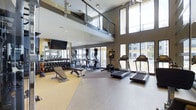This two-story fitness center offers a well-appointed and thoughtfully designed workout environment that caters to a wide range of exercise preferences. The lower level serves as the main gym floor, featuring an open layout with high ceilings that create a spacious, energizing atmosphere. Large floor-to-ceiling windows line the walls, flooding the space with abundant natural light and providing views of the surrounding exterior. The main floor is equipped with a comprehensive selection of cardio machines, including treadmills and elliptical trainers, as well as a dedicated free weights area with a full dumbbell rack, adjustable benches, and a wall-mounted flat-screen television. Flooring transitions between hardwood-style and rubberized mat surfaces, delineating cardio and strength training zones. A sleek steel cable railing system defines the mezzanine level above, which is visible from the main floor and accessible via the second level. The upper floor houses a cycling studio with multiple stationary and spin bikes, benefiting from its own set of large windows that bring in natural light. Recessed and track lighting throughout both levels ensures excellent visibility at any time of day. The overall design aesthetic is contemporary and clean, with neutral tones, modern finishes, and an open, airy feel that makes the facility both functional and inviting.