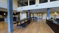This contemporary amenity space is designed with an open-concept layout that maximizes its generous square footage. The most striking architectural feature is the dramatic double-height ceiling, accented by large upper-level windows that flood the interior with natural light and create an airy, expansive atmosphere.

The space is organized into distinct functional zones while maintaining a seamless flow throughout. A fully equipped kitchen anchors one end, featuring stainless steel appliances, a built-in refrigerator, and a bar-style counter with seating — ideal for hosting and social gatherings. Adjacent to the kitchen, a long dark-toned dining table with matching bench seating provides ample room for group meals or collaborative use.

At the center of the room, a striking double-sided fireplace with brick detailing serves as a bold architectural focal point, separating the dining area from the recreation zone. The recreation area includes a full-sized pool table, perfect for leisure and entertainment. Glass-paneled double doors at the far end offer additional connectivity to adjacent spaces.

Warm hardwood-style flooring runs throughout, complementing the contemporary dark furnishings and neutral white walls, resulting in a polished, inviting communal environment.