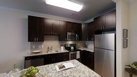 This single-floor kitchen showcases a cohesive design centered around rich, dark espresso-toned cabinetry that extends from floor to ceiling, providing generous storage throughout. The upper and lower cabinets feature sleek bar-pull hardware, complemented by elegant crown molding that adds a refined finishing touch along the ceiling line.

The countertops are finished in light granite with subtle veining, offering a striking contrast against the dark cabinetry. A matching granite kitchen island provides additional prep space and casual use. The backsplash features a neutral tile in a subtle linear pattern, tying the design elements together cohesively.

Stainless steel appliances include a French-door refrigerator, an electric range with a built-in microwave above, and a dishwasher, delivering a modern and functional cooking environment. The undermount sink is positioned along the perimeter counters, accompanied by a contemporary faucet.

Overhead, a large flush-mount fluorescent light fixture ensures bright, even illumination across the workspace. Crown molding details reinforce the polished, well-finished character of this thoughtfully designed kitchen space.