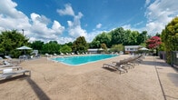 Located at 4825 Commercial Plaza St in Winston-Salem, North Carolina, this outdoor pool area offers a well-appointed recreational amenity designed for relaxation and leisure. The centerpiece is a generously sized rectangular swimming pool with clear, blue-tinted water, marked with depth indicators and equipped with sturdy entry railings for safe access. Surrounding the pool is a broad, paved sundeck that provides ample space for movement and socializing. Numerous reclining lounge chairs are arranged around the perimeter of the deck, offering residents plenty of options for sunbathing and unwinding. A shaded umbrella station provides a comfortable retreat from direct sun. The pool area is fully enclosed by a chain-link fence, ensuring privacy and security. Mature trees — including a striking magnolia — frame the space and provide a natural, verdant backdrop that enhances the outdoor ambiance. Ornamental shrubs and plantings along the fence line add additional greenery and visual interest. Floodlighting is present for extended evening use. The surrounding community buildings, constructed of brick and neutral tones, are visible beyond the enclosure, reinforcing the residential character of the setting. Overall, the pool area presents a clean, functional, and inviting outdoor retreat well-suited for warm-weather enjoyment.