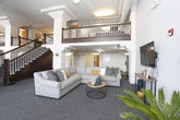 This inviting communal space blends industrial character with modern comfort in a thoughtfully designed open-plan layout. Soaring ceilings with exposed beams and ductwork lend the space an airy, loft-like feel, while a mezzanine balcony with a wood-railed staircase adds vertical drama and architectural interest.

The main floor is organized into distinct yet connected zones. A generously sized lounge area anchors one end of the space, featuring a large sectional sofa in soft grey accented with mustard and neutral throw pillows, arranged around a round wire-and-wood coffee table — a nod to the industrial aesthetic throughout. A wall-mounted flat-screen television completes this relaxed gathering spot.

Adjacent to the lounge, a pair of mid-century-style armchairs sit atop a woven area rug, framed by decorative wall art and a warm floor lamp, creating a quieter reading or conversation nook. A dining area nearby offers a dark wood table and chairs set beside large, multi-pane factory-style windows that flood the space with natural light and offer views of the surrounding neighborhood.

The recreation area features a full-sized pool table bathed in natural light from expansive windows, accompanied by a bench and side table for a comfortable retreat. Throughout, neutral grey carpet, white walls, and structural columns tie the space together into a cohesive, welcoming environment.